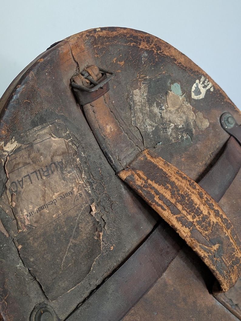 Pu&ograve; includere: Una cappelliera in pelle marrone usurata con manico logoro, fibbia in metallo e due etichette di carta strappate. Un'etichetta mostra chiaramente il testo AURILLAC e Gare destinataire, l'altra mostra testo parziale incluso GES PL.