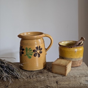 May include: A yellow ceramic pitcher with a handle and dark brown and green floral patterns, a yellow and brown ceramic pot with a wooden pestle, and a square block of light brown soap, accompanied by a small bundle of dried lavender sprigs.