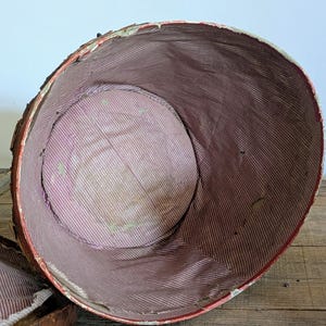 Pu&ograve; includere: Un contenitore rotondo aperto superiormente con un bordo esterno rosso e una superficie esterna testurizzata simile a corteccia chiara. L'interno &egrave; foderato con un tessuto a righe rosa e bianche, che mostra segni di usura e piccoli strappi.