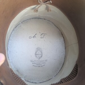 Pu&ograve; includere: L'interno di un cappello a cilindro con una fodera chiara e le iniziali A.D. stampate in nero. Un logo circolare riporta BERTEIL, 18, RUE DE L'ANC. COMEDIE PARIS e MEDAILLES D'OR 1878-1889. Un piccolo fiocco in tessuto beige &egrave; fissato in alto.