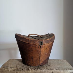 Pu&ograve; includere: Una cappelliera in pelle marrone usurata con coperchio curvo, maniglia centrale per il trasporto e chiusura a scatto in metallo. Il contenitore presenta cuciture visibili lungo i lati e una texture invecchiata su tutta la superficie esterna.