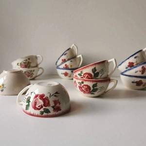 Könnte beinhalten: Eine Sammlung von Vintage-Teetassen und Untertassen aus Keramik. Die Tassen weisen florale Designs in Rot, Blau und Gelb auf, mit weißen Hintergründen und gebogenen Griffen. Einige Tassen haben einen roten oder blauen Rand. Die Untertassen sind ebenfalls mit Blumen verziert.