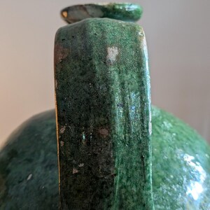 May include: Close-up of a green ceramic jug with a handle and a small spout. The jug has a textured surface with visible imperfections and a glossy finish. The handle is rectangular and the spout is rounded.