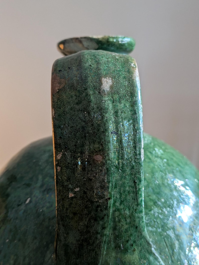 May include: Close-up of a green ceramic jug with a handle and a small spout. The jug has a textured surface with visible imperfections and a glossy finish. The handle is rectangular and the spout is rounded.