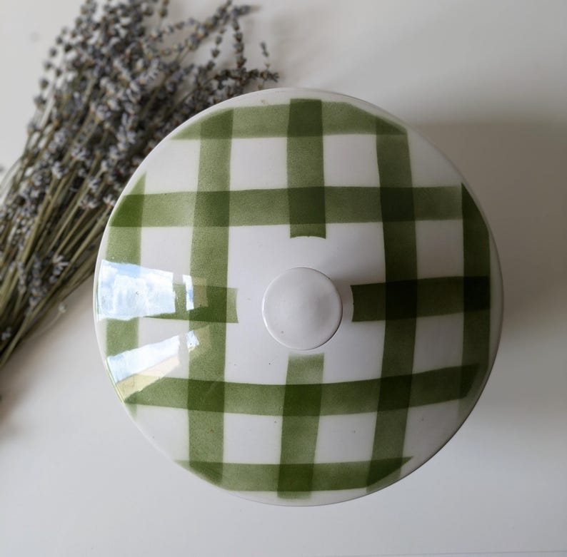 May include: A white ceramic lid with a green plaid pattern. The lid has a small, round, white knob in the center. The design is reminiscent of a picnic blanket. Dried lavender is visible in the background.