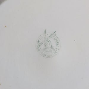 May include: White ceramic plate with a faded green stamp in the center. The stamp features a windmill design with the words "Moulin des Loups France" encircling the image. The plate has a slightly curved edge.