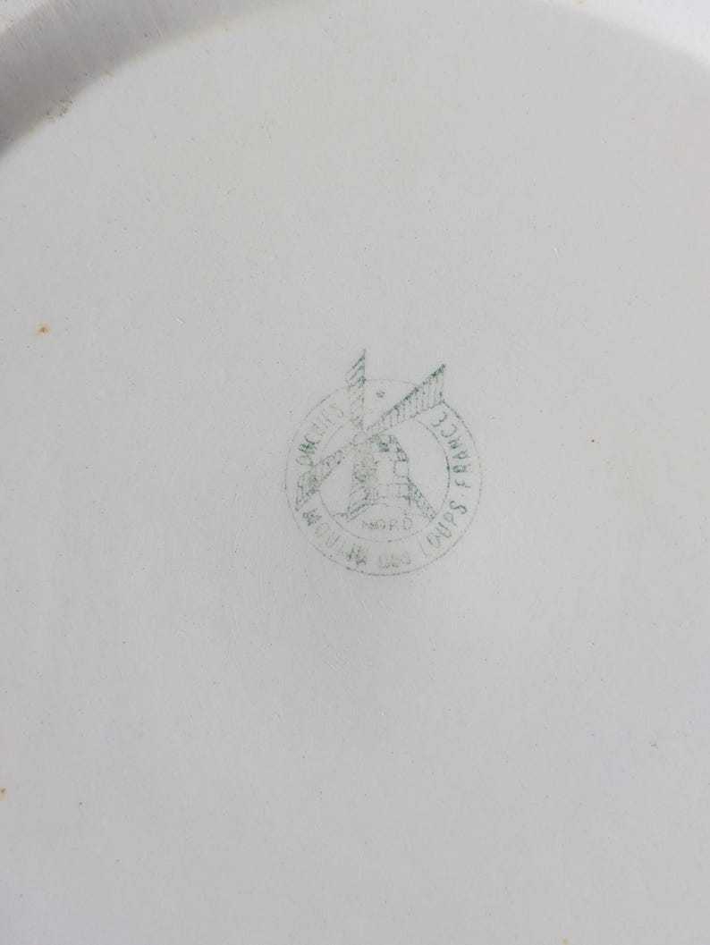 May include: White ceramic plate with a faded green stamp in the center. The stamp features a windmill design with the words "Moulin des Loups France" encircling the image. The plate has a slightly curved edge.