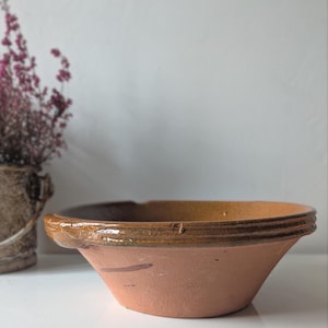 May include: A terracotta-colored bowl with a wide rim and a slightly flared shape. The rim has a darker brown band with horizontal ridges. The interior of the bowl has a golden-brown hue. The bowl is set against a white background.