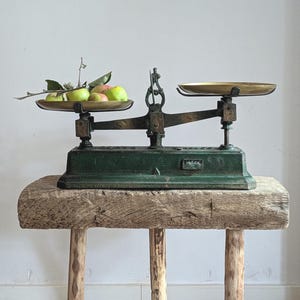Peut inclure: Une balance à plateaux ancienne avec une base en fonte vert foncé et deux plateaux en laiton. Le plateau de gauche contient plusieurs pommes vertes et rouges avec des tiges et des feuilles. La balance possède un fléau central vertical.