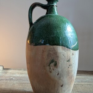 May include: A large, antique stoneware jug with a green glazed top and a natural, unglazed lower half. The jug features a curved handle and a rounded body, showcasing a rustic, aged aesthetic. The jug is likely used for storing liquids.