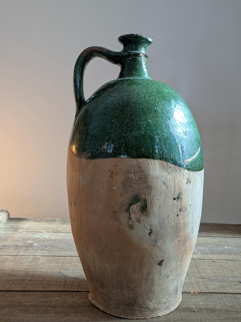 May include: A large, antique stoneware jug with a green glazed top and a natural, unglazed lower half. The jug features a curved handle and a rounded body, showcasing a rustic, aged aesthetic. The jug is likely used for storing liquids.