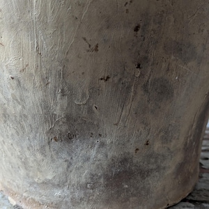 May include: Close-up of a weathered, beige-colored ceramic pot. The pot has a textured surface with visible brushstrokes and dark spots. The base of the pot is visible, showing a slightly uneven edge.