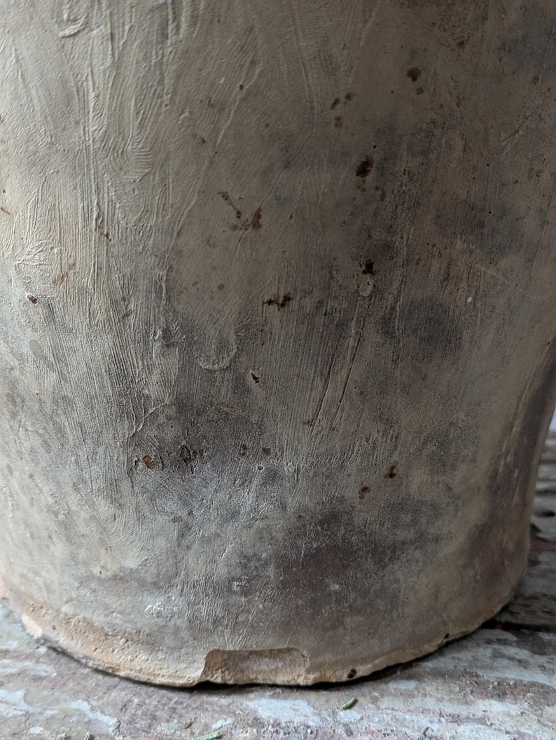May include: Close-up of a weathered, beige-colored ceramic pot. The pot has a textured surface with visible brushstrokes and dark spots. The base of the pot is visible, showing a slightly uneven edge.