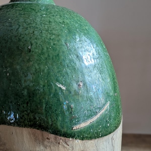 May include: A vintage green glazed ceramic jug with a rounded body and a narrow neck. The upper portion is a mottled green, while the lower section is a lighter, unglazed color. The jug has a rustic, aged appearance.
