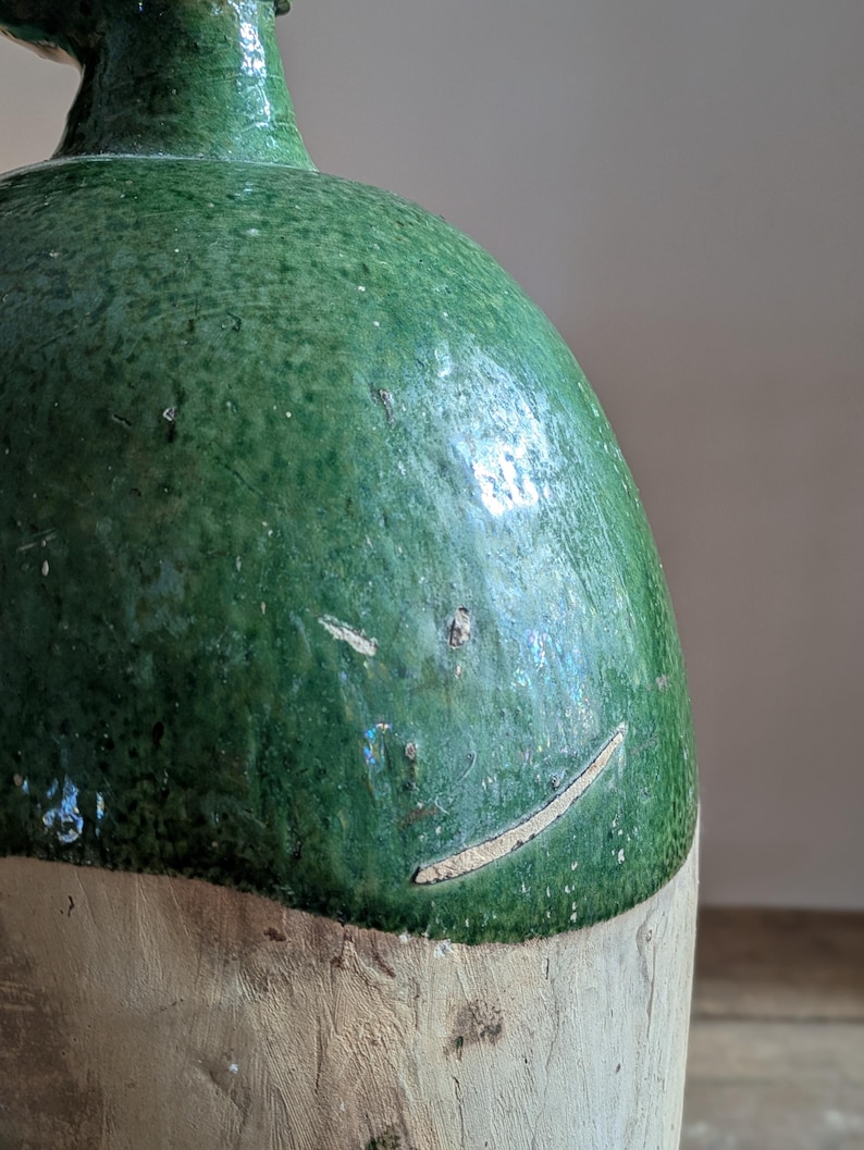 May include: A vintage green glazed ceramic jug with a rounded body and a narrow neck. The upper portion is a mottled green, while the lower section is a lighter, unglazed color. The jug has a rustic, aged appearance.
