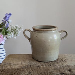 Puede incluir: Una jarra de cerámica de gres gris claro con dos asas curvas. La jarra tiene un borde más oscuro y está sobre una superficie de madera envejecida. Un jarrón de flores está a la izquierda de la jarra. Estilo rústico.