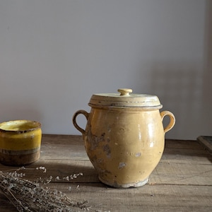 Könnte beinhalten: Ein gelber Keramiktopf mit Deckel und zwei Griffen, neben einem kleineren gelben und braunen Keramikbehälter. Der Topf hat ein abgenutztes Aussehen. Die Gegenstände stehen auf einer Holzoberfläche.