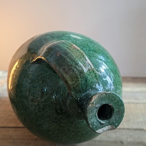 May include: A green glazed ceramic jug with a rounded body and a curved handle. The jug has a dark green color with lighter green highlights. The opening is circular. The jug is sitting on a wooden surface.