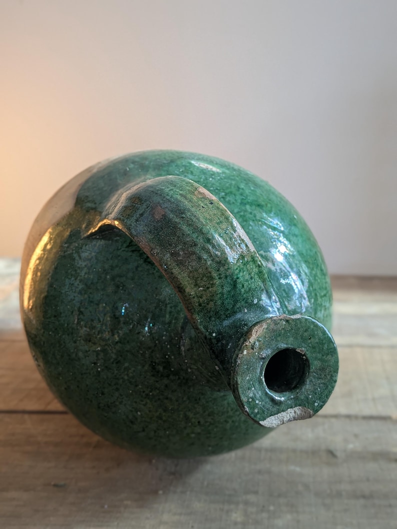 May include: A green glazed ceramic jug with a rounded body and a curved handle. The jug has a dark green color with lighter green highlights. The opening is circular. The jug is sitting on a wooden surface.