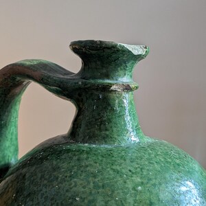May include: A close-up of a green ceramic jug with a handle. The jug has a rounded body and a narrow neck with a spout. The handle curves from the neck to the side of the jug. The surface has a textured appearance.