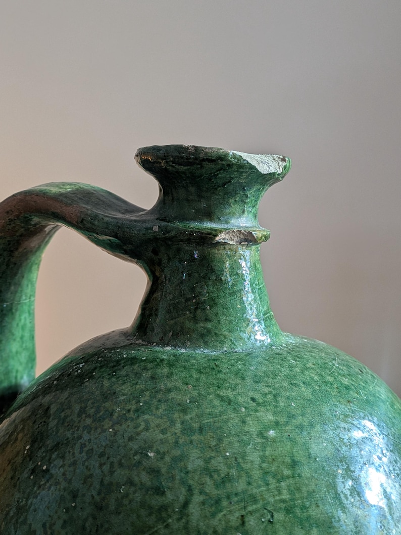 May include: A close-up of a green ceramic jug with a handle. The jug has a rounded body and a narrow neck with a spout. The handle curves from the neck to the side of the jug. The surface has a textured appearance.