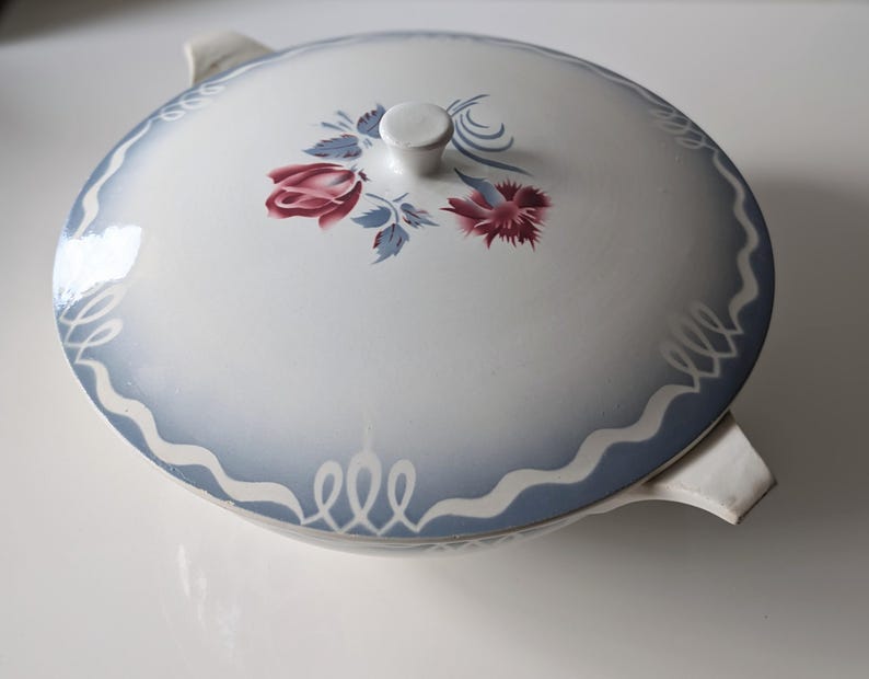 Puede incluir: Un plato de servir de cer&aacute;mica blanca con un dise&ntilde;o floral azul y blanco. El plato tiene dos asas y una tapa.