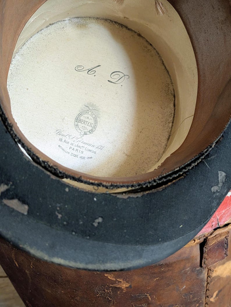Pu&ograve; includere: Un cilindro con una fascia esterna nera e una fodera interna di colore chiaro. L'interno presenta le iniziali A. D. e un'etichetta stampata con la scritta Manufacture de Chapeaux Berteil, 18 Rue de Lang Comedie Paris, Medailles Dor 1878-1889.