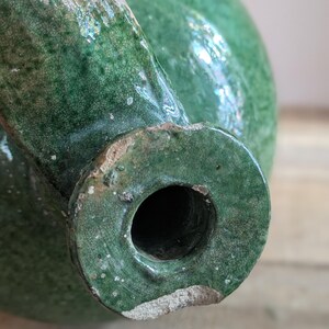 May include: Close-up of a green ceramic pitcher with a spout and a round opening. The pitcher has a crackled glaze and a distressed appearance. The ceramic is a deep, rich green color.