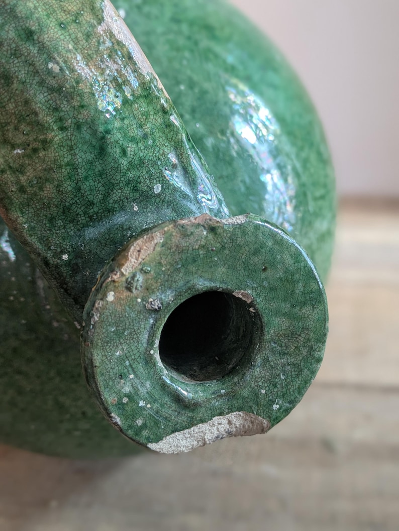 May include: Close-up of a green ceramic pitcher with a spout and a round opening. The pitcher has a crackled glaze and a distressed appearance. The ceramic is a deep, rich green color.
