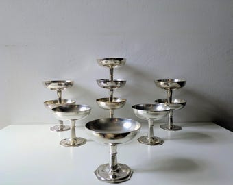 Può includere: Una collezione di sette coppe in metallo color argento disposte in formazione a gradini. Ogni coppa presenta una coppa larga e poco profonda, uno stelo sottile e una base circolare. La superficie metallica riflette la luce, creando riflessi.