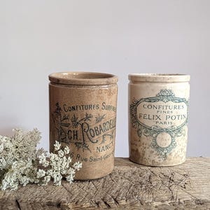 May include: Two cylindrical stoneware crocks. The left crock is tan with black text reading Confitures Surfinies Ch. Robardelle Nancy 26 Rue Saint-Georges. The right crock is cream with a speckled finish and green text reading Confitures Fines Félix Potin Paris.