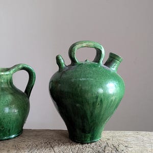May include: A green glazed ceramic vessel with a rounded body, a central arched handle, a narrow spout on the left, and a wider opening on the right. A partial view of a second similar green ceramic pitcher is visible on the left side of the frame.