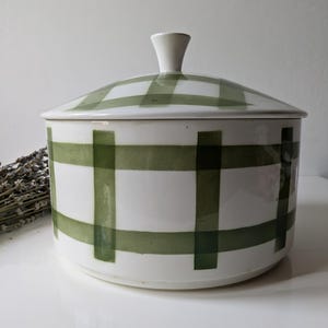 May include: A white ceramic lidded container with a green plaid pattern. The lid has a small, white handle. The container is round and sits on a white surface. Dried lavender is visible to the left of the container.