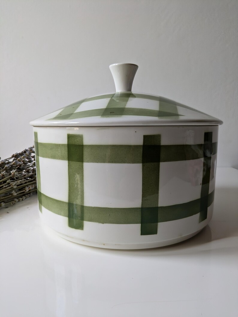 May include: A white ceramic lidded container with a green plaid pattern. The lid has a small, white handle. The container is round and sits on a white surface. Dried lavender is visible to the left of the container.