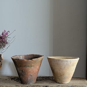 Puede incluir: Dos macetas de terracota cónicas y desgastadas, con bordes ásperos e irregulares. Una es de color marrón rojizo, la otra de color beige claro. Se encuentran sobre una superficie de madera rústica, sugiriendo una estética vintage o antigua. Medidas en centímetros.