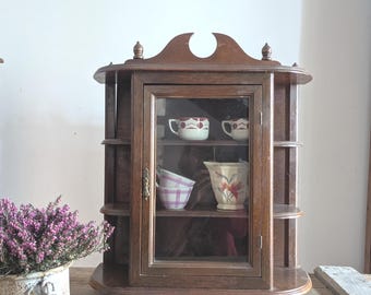 May include: A vintage wooden display cabinet with a glass door, showcasing decorative cups and a pitcher. The cabinet is dark brown with rounded shelves and a decorative top. A pot of purple flowers sits to the left on a wooden surface.
