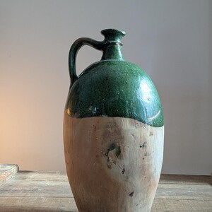 May include: A large, antique stoneware jug with a green glazed top and a natural, unglazed lower half. The jug has a handle and a rounded body, showcasing a rustic, aged appearance. The jug is displayed on a wooden surface.