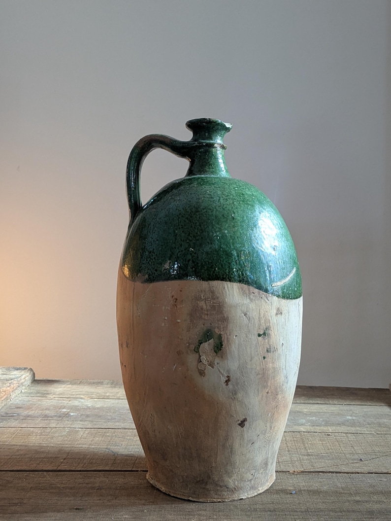 May include: A large, antique stoneware jug with a green glazed top and a natural, unglazed lower half. The jug has a handle and a rounded body, showcasing a rustic, aged appearance. The jug is displayed on a wooden surface.