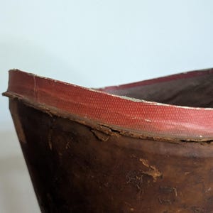 Pu&ograve; includere: Uno stivale in pelle marrone usurata con un bordo rosso testurizzato lungo la parte superiore. La pelle mostra segni di usura con crepe visibili e una consistenza invecchiata, mentre la fodera interna appare scura e a coste.