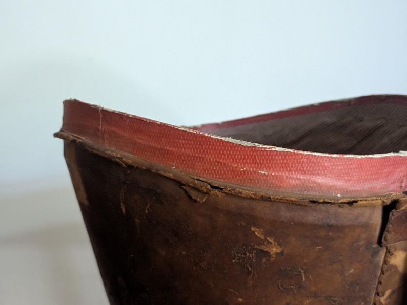 Pu&ograve; includere: Uno stivale in pelle marrone usurata con un bordo rosso testurizzato lungo la parte superiore. La pelle mostra segni di usura con crepe visibili e una consistenza invecchiata, mentre la fodera interna appare scura e a coste.