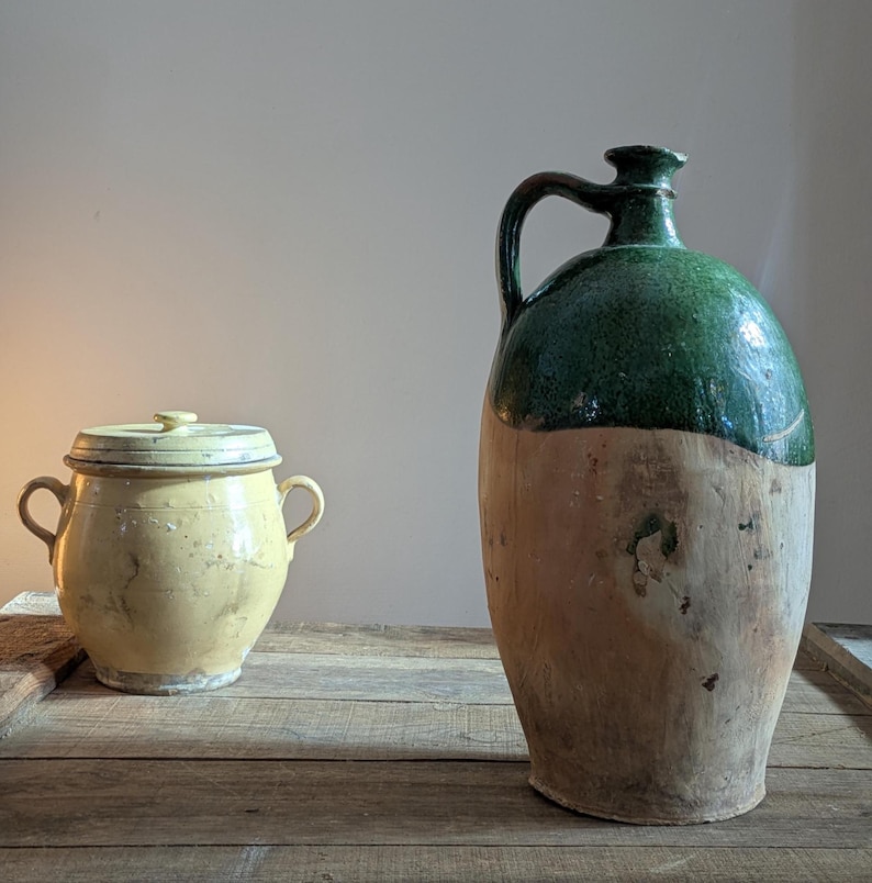 May include: Two antique ceramic vessels on a wooden surface. A small, yellow lidded pot with handles sits next to a large, green-topped jug with a handle. The jug has a light brown base. The items appear aged and rustic.