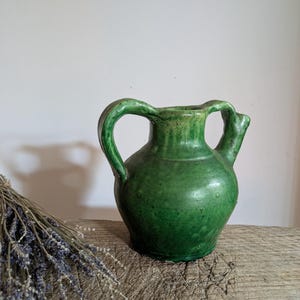 May include: A green ceramic pitcher with a rounded body, a wide mouth, and two symmetrical handles on either side. The surface features a glossy, mottled green glaze with subtle texture and small bumps throughout the exterior of the vessel.