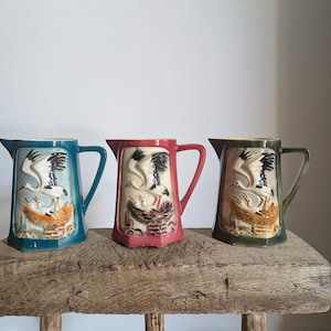 May include: Three ceramic jugs featuring a relief design of a stork standing over a nest with chicks. The jugs are coloured in teal, red, and dark green, each with a handle and a spout. The relief artwork shows the birds in white and black.
