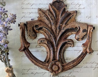May include: A cast iron door knocker featuring a decorative acanthus leaf design with a distressed, rusted brown finish. The hardware includes a central vertical leaf element and a hinged, curved ring pull attached to the sides of the base.