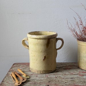 May include: A cylindrical cream-coloured ceramic crock with two side handles and a thick rim. Beside it lie three small wooden pastry crimpers with serrated wheels. The crock has a smooth, glazed finish with subtle brown speckles and wear.