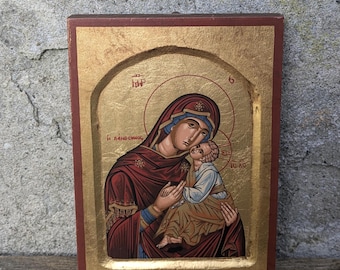 Può includere: Un'icona in legno dipinta a mano che rappresenta la Vergine Maria che tiene in braccio il bambino Gesù. L'icona è incorniciata in oro e rosso.