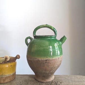 Puede incluir: Una jarra de cerámica esmaltada en verde con cuerpo redondeado, asa superior, asa lateral y un pequeño pico vertedor. La parte superior tiene un acabado de esmalte verde brillante, mientras que la base permanece en arcilla marrón natural.