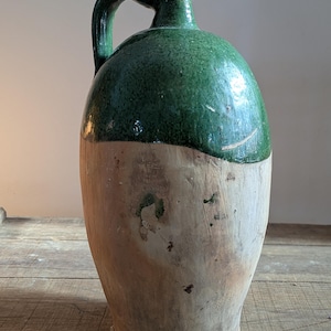 May include: A large, antique ceramic jug with a handle. The top half is glazed in a vibrant green, while the bottom half is a natural, unglazed tan color. The jug has a rounded body and a narrow neck.