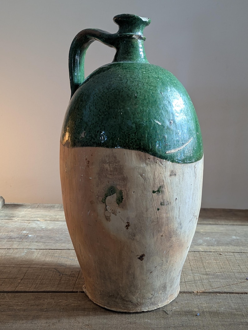 May include: A large, antique ceramic jug with a handle. The top half is glazed in a vibrant green, while the bottom half is a natural, unglazed tan color. The jug has a rounded body and a narrow neck.