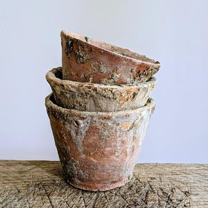 May include: A stack of three terracotta plant pots with a weathered, textured surface. The pots feature a reddish-brown clay base with patches of grey and white mineral deposits, showing a rough, aged appearance on the exterior rims and sides.